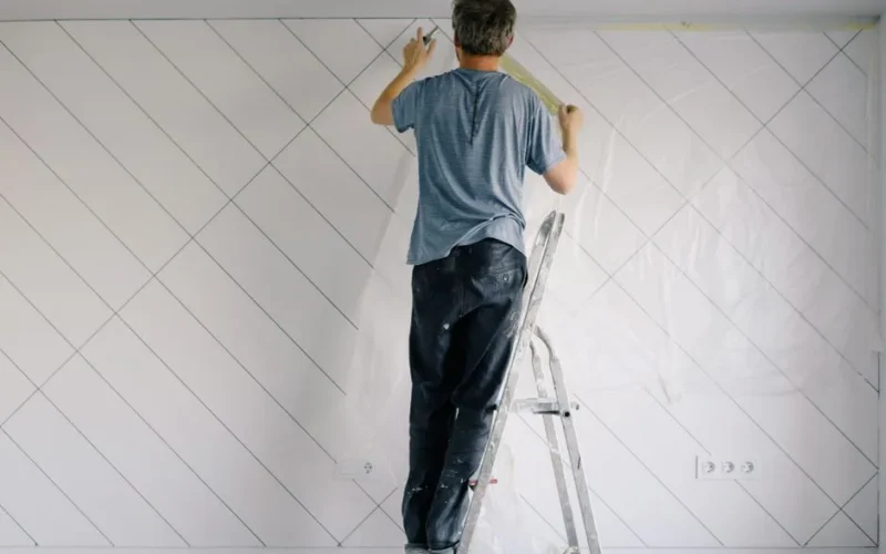 Full body back view of faceless male in shirt and dirty pants repainting wall while standing on ladder in bright empty room in daytime
