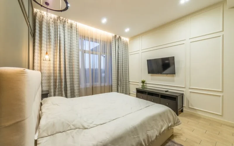 Interior of modern bedroom with comfortable bed and big window covered with curtains under luminous lights