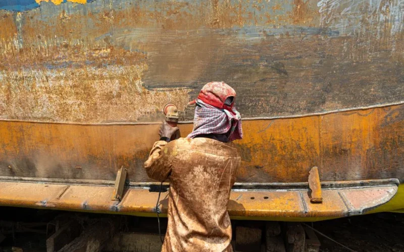A worker sands a rusty metal surface outdoors using a power tool, wearing protective clothing.