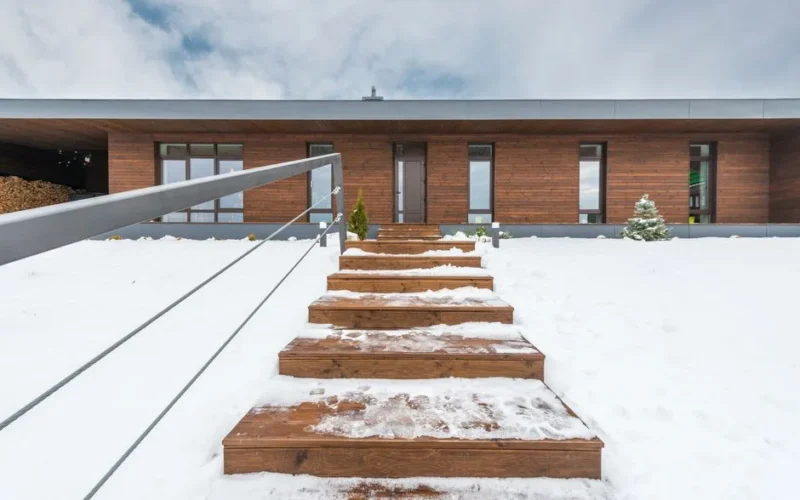 A contemporary wooden house with snow-covered steps leading to the entrance, captured in a winter setting.