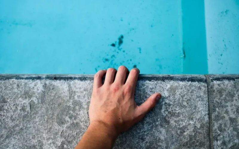 Close-up of a human hand on a concrete edge by a pool with clear blue water.