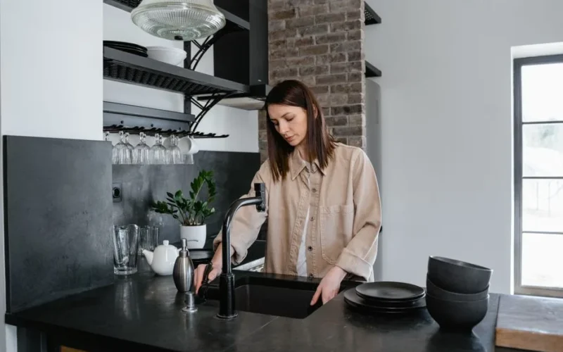 A young woman stands by a sink in a modern kitchen, washing dishes with natural light pouring in.