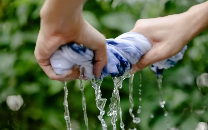 Hands of faceless person with wet dyed fabric in hands with waterdops standing near green plants on blurred background in countryside