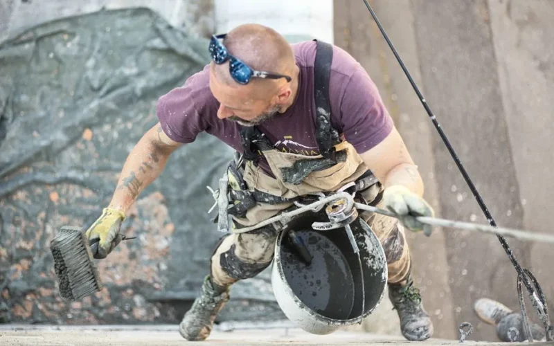 From above of man in safety harness hanging above ground and smoothing wall skim coat with liquid plaster using coarse brush