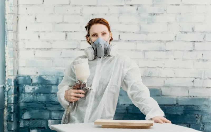 Woman in protective suit and gas mask using spray gun indoors against a painted brick wall.