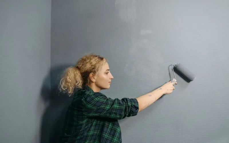 Young woman uses a paint roller to apply grey paint on an indoor wall, renovating her space.