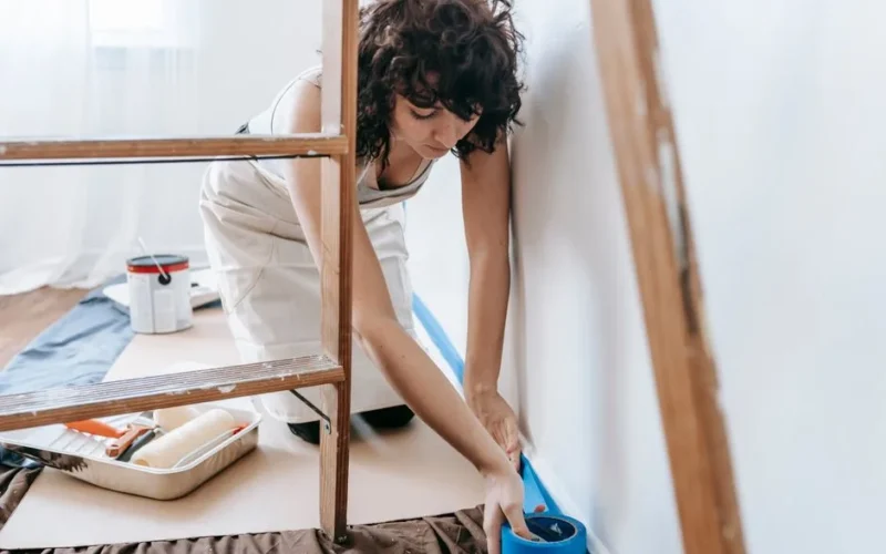 Woman uses tape to prep wall for painting, part of a DIY home renovation project indoors.