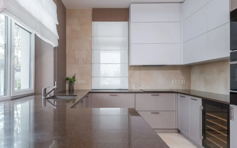 Minimalist interior kitchen design with stone countertop of marble color with sink and chrome plated faucet lying on cupboard with built in stove and electric appliances in daylight