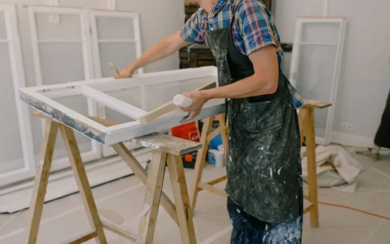 Unrecognizable male master wearing dirty apron standing near window with white frames on wooden workbench while working in professional studio