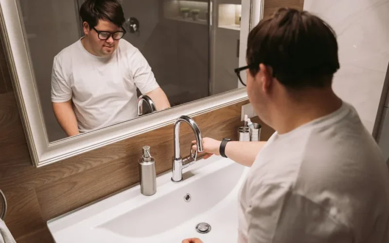 A man with Down syndrome using a bathroom sink while wearing glasses.