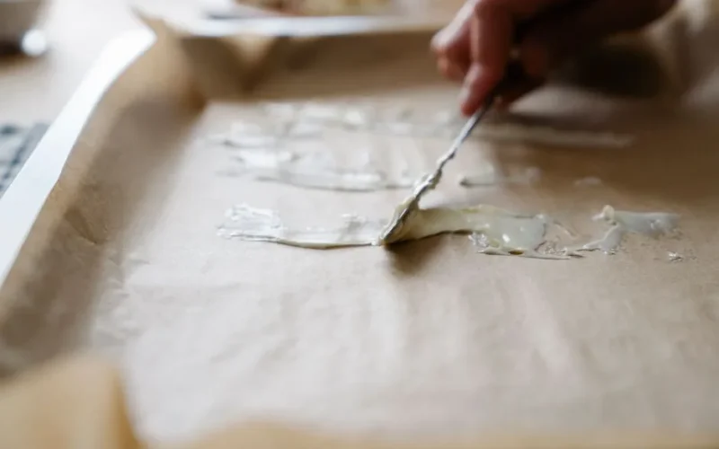 Close-up of hand spreading butter on baking tray lined with parchment paper, preparing for baking.