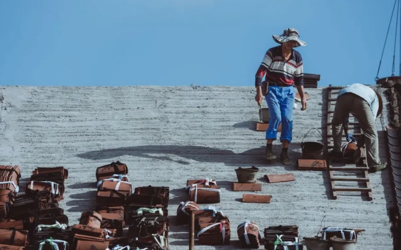Two construction workers installing roof tiles on a building under a clear blue sky.