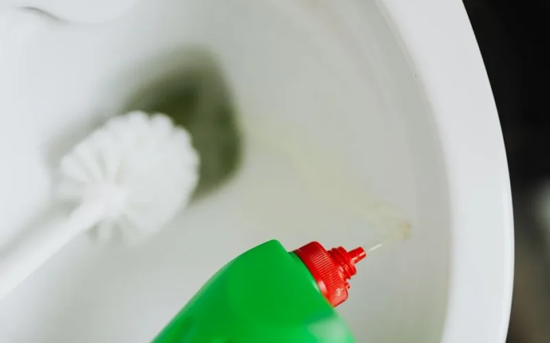 Top view of liquid detergent pouring from green plastic bottle in toilet bowl with toilet brush