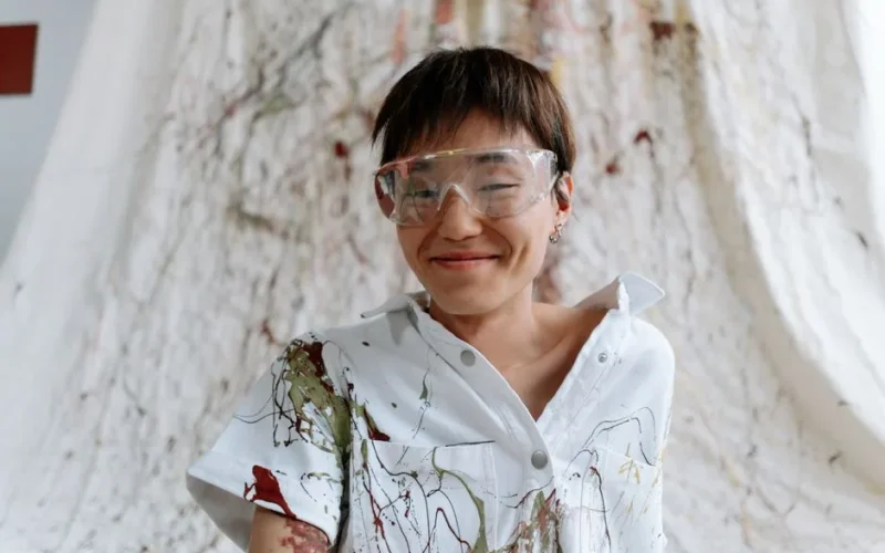 A cheerful Asian artist with short hair in safety glasses and a paint-splattered white shirt.