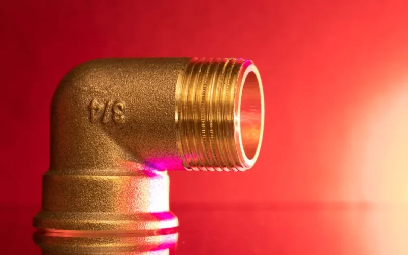 Close-up of a shiny brass pipe elbow fitting against a vibrant red backdrop, showcasing industrial elegance.