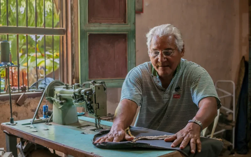 The image shows an elderly man with Hispanic features, sitting in front of the window of his bahareque house, a type of traditional construction in Venezuela made of mud and other natural materials. The soft daylight enters through the window, illuminating the makeshift workspace where the man, with a calm and focused expression, dedicates himself to sewing the upholstery of his car. The sewing machine, an old Singer model, stands out in the foreground, with its classic design and handwheel that has been adapted with a washing machine motor under the table. The upholstery that the man is repairing shows signs of wear but also care, symbolizing effort and dedication in times of scarcity. In the background, the bahareque walls, with their earthy texture and marks of the passage of time, speak of the resilience of the structures and the people who inhabit them. This photograph, taken a decade ago, is a testament to hope and human tenacity at a time when self-management became an everyday response to economic adversity. More than a simple capture of a moment in an individual's life, it reflects the shared history of a community that, even in difficult times, clings to the hope for better days.