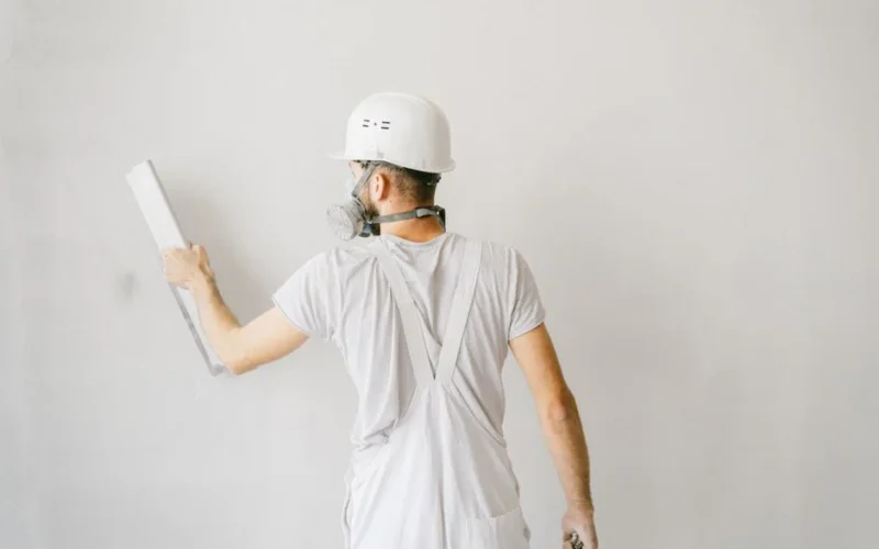 Back view of a construction worker in safety gear plastering a white wall.
