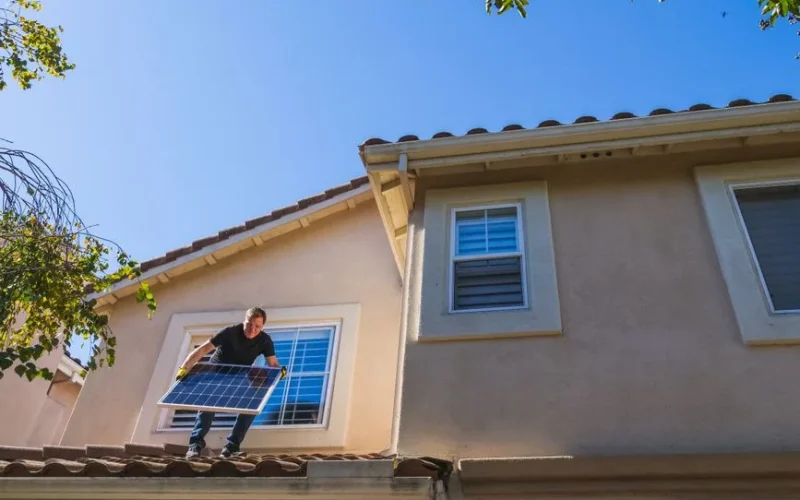 Man installing a solar panel on a rooftop under a clear blue sky, promoting renewable energy.
