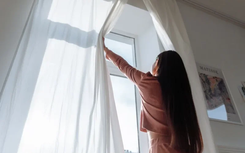 Woman opens white curtains in bright room, revealing a sunny day outside.