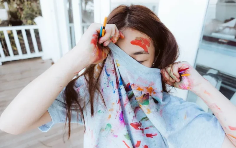 A playful artist covered in vibrant watercolors, hiding face with a colorful shirt.