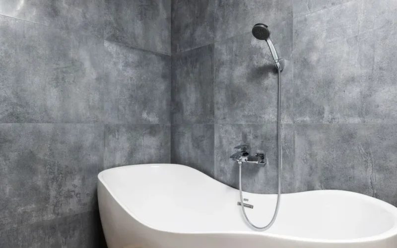 Interior of light modern bathroom with white bath under shower on gray tile wall
