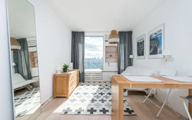 Wooden table and drawer placed in room with big window and wall decorated with pictures in light room with mirror